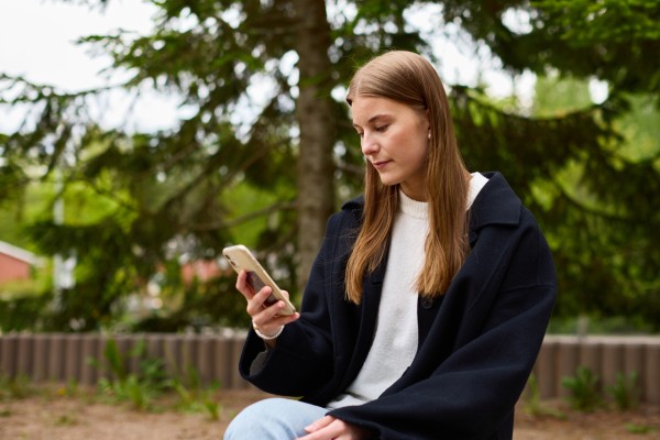 Nuori nainen katsoo puhelintaan. Taustalla vihreitä puita.