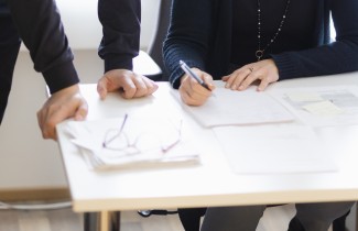 Kahdet kädet nojaavat pöytään, toinen henkilö kirjoittaa kynällä paperille.