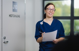 Ruskeatukkainen silmälasipäinen hoitaja hymyilee. Hänellä on kädessään paperi ja kynä.