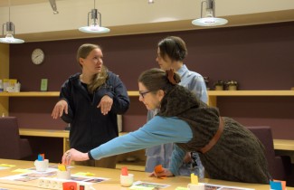 Villasukkakilpailun raatiin kuuluvat Tuire Selin ja Kati Kallimo keskustelevat kilpailutöistä. Kristiina Lempiäinen-Trzaska kurottaa pöydän yli sujauttamaan pisteytyslapun yhdelle kilpailutyölle.