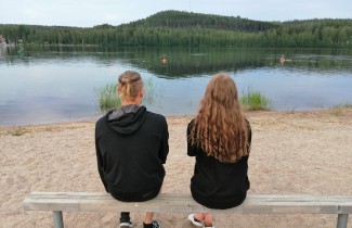 Kaksi henkilöä istuu puisella kapealla penkillä selin kameraan järven rannassa