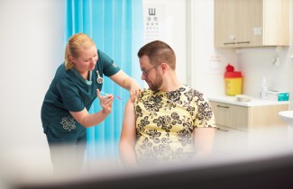 Kuvassa on keltapaitainen aikuinen miesoletettu henkilö, jota rokottaa naisoletettu terveydenhuollon ammattilainen.