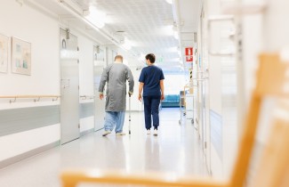 Ikäihminen keppeineen laitoksen käytävällä hoitajan saattamana