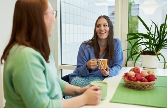 Kaksi henkilöä istuu pöydän ääressä vieretysten mukit kädessään hymyillen toisilleen.