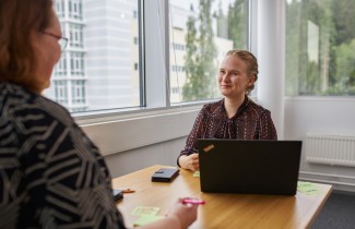 Kuvassa työtoverit juttelevat toistensa kanssa puisen pöydän ääressä. Pöydällä on auki kannettava tietokone.