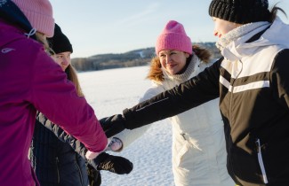 Neljä hymyilevää ihmistä on kokoontunut joukkuetervehdykseen talvimaisemassa. He ovat yhdistäneet kätensä ringin keskelle, valmiina yhteiseen kannustushuutoon.