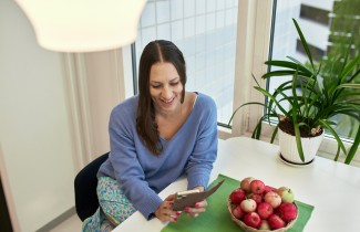 Henkilö puhelimella pöydän ääressä, omenakulho ja viherkasvi taustalla.