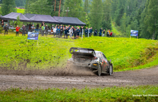 Ralliauto ajaa hiekkatietä, katsojia kauempana.
