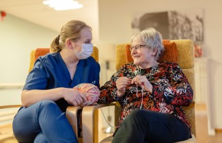 Hoitaja ja ikääntynyt nainen istuvat iloisina vierekkäin nojatuoleilla toisiaan katsoen.