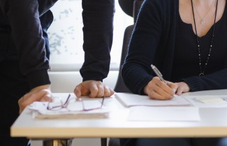 Pöydällä on papereita ja silmälasit, yhden henkilön kädet nojaavat pöytää vasten ja toinen henkilö kirjoittaa paperille. 