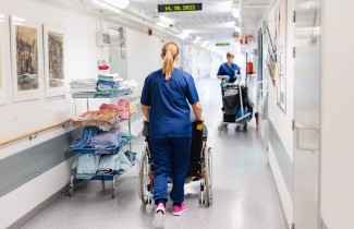 Hoitaja kuljettaa pyörätuolissa istuvaa potilasta terveyskeskuksen käytävällä. Käytävän varrella on siivouskärry ja siivooja.