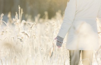 Ulkovaatteisiin pukeutunut aikuinen seisoo selin talvisessa peltomaisemassa. Aurinko paistaa. 