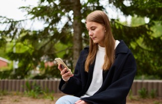 Nuori nainen katsoo puhelintaan. Taustalla vihreitä puita.