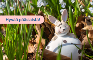 Pääsiäispupukoriste istuu maasta pilkottavien kukanvarsien seassa. Kuvassa on teksti 'Hyvää pääsiäistä!'.   