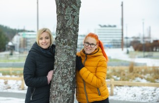 Kaksi naista nojaa puuhun eri puolilta alkukeväisessä kaupunkimaisemassa.