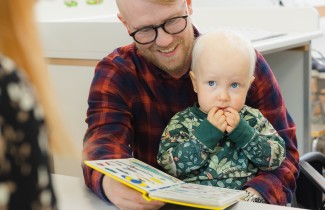 Lapsi istuu isän sylissä, isä hymyilee ja lukee lapselle kuvakirjaa.