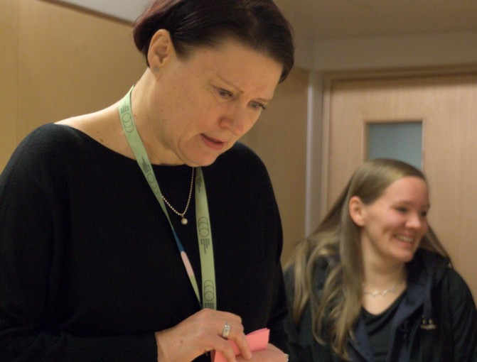 Raatiin kuuluva Nina Peränen tutkii tarkkaan kilpailutöitä miettiessään pisteytystä. Taustalla Tuire Selin hymyilee ja pukee takkia ylleen - hän on saanut omat pisteensä jaettua.