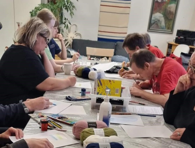 Ihmisiä on kerääntynyt pitkän pöydän ympärille piirtämään ja ideoimaan sukkamalleja. Pöydällä on kyniä, papereita ja monta lankakerää.