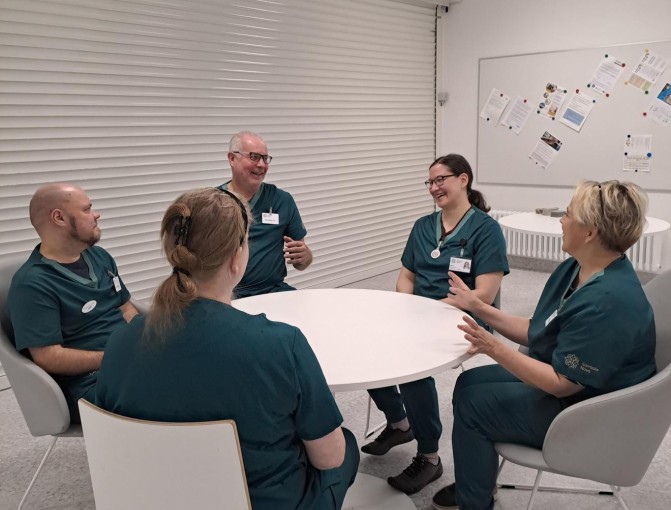 Viisi hymyilevää ammattilaista ovat kokoontuneet pyöreän pöydän ääreen keskustelemaan perushoidon kehittämisestä.