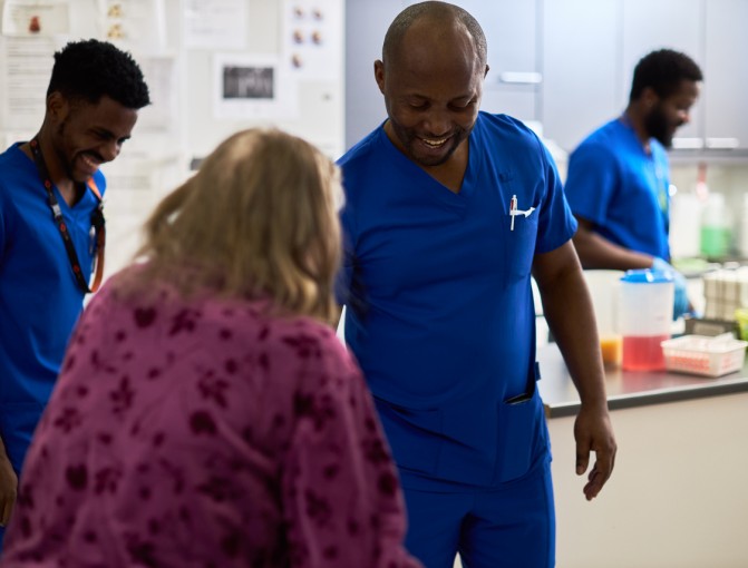 Sairaanhoitaja Fidele kohtaa hymyillen ikääntyneen asiakkaan. Taustalla muutama hymyilevä hoitajakollega valmistelee ruokailua.
