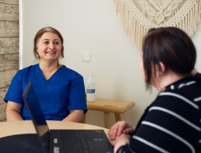 Hoiva-avustaja Seher istuu hymyillen pöydän ääressä palveluvastaava Reea Mäkisen kanssa.