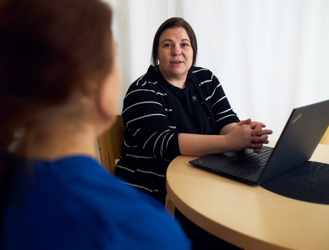 Palveluvastaava Reea Mäkinen istuu pöydän ääressä kädet ristissä kannettava tietokone edessään ja katsoo hoiva-avustaja Seheriä, joka istuu vastapäätä.