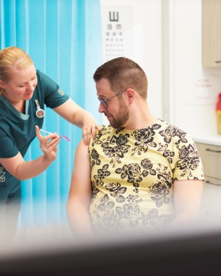 Kuvassa on keltapaitainen aikuinen miesoletettu henkilö, jota rokottaa naisoletettu terveydenhuollon ammattilainen.