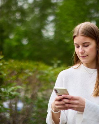 Nuori nainen katsoo puhelintaan. Taustalla puita ja pensaita.