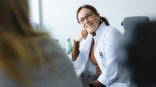 Lääkäri istuu tuolissa nojaten päätä käteensä ja katsoo hymyilleen potilasta.