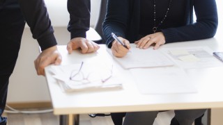 Kahdet kädet nojaavat pöytään, toinen henkilö kirjoittaa kynällä paperille.