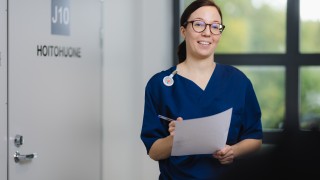 Ruskeatukkainen silmälasipäinen hoitaja hymyilee. Hänellä on kädessään paperi ja kynä.