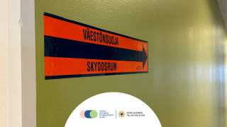 Väestönsuojan opastetarra rakennuksen seinällä