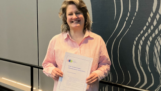 Henkilö hymyilee kameralle ja pitää molemmin käsin kiinni A4 kokoisesta paperista, jossa lukee palkinnon nimi.