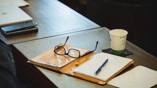 Pöydällä silmälasit, avattu vihko, kynä ja pahvimuki