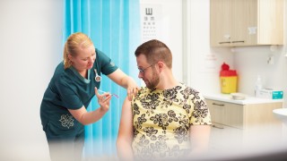 Kuvassa on keltapaitainen aikuinen miesoletettu henkilö, jota rokottaa naisoletettu terveydenhuollon ammattilainen.