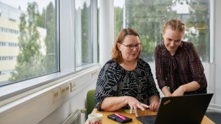 Kaksi työntekijä istuvat yhteisen tietokoneen äärellä ja katselevan tietokoneen näytölle.