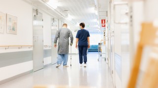 Ikäihminen keppeineen laitoksen käytävällä hoitajan saattamana