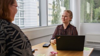 Kuvassa työtoverit juttelevat toistensa kanssa puisen pöydän ääressä. Pöydällä on auki kannettava tietokone.