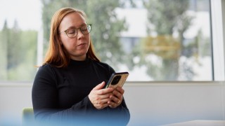 Kuvassa aikuinen naisoletettu katsoo kännykkäänsä. Naisella on pitkät, punertavat hiukset ja musta poolupaita. Nainen istuu istuu tuolilla pöydän ääressä. Takana olevasta ikkunasta näkyy metsämaisema.