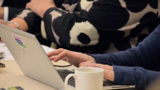 Kuvassa on tietokoneella kirjoittavat kädet. Koneen vieressä on valkoinen kahvikuppi.