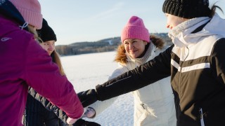 Neljä hymyilevää ihmistä on kokoontunut joukkuetervehdykseen talvimaisemassa. He ovat yhdistäneet kätensä ringin keskelle, valmiina yhteiseen kannustushuutoon.