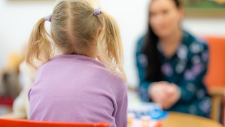 Lapsi istuu selin ja katsoo taustalla olevaa aikuista