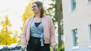 Hymyilevä nainen seisoo kyynärsauvojen kanssa parkkipaikalla aurinkoisessa syyssäässä