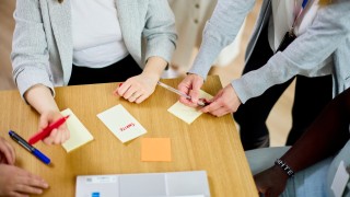 Pöydällä on post-it-lappuja, joista yhdessä lukee tavoite. Pöydän ympärillä on neljä ihmistä, joiden kasvot eivät näy.