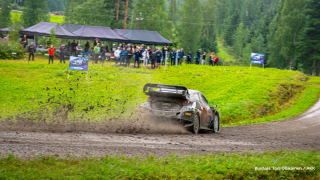 Ralliauto ajaa hiekkatietä, katsojia kauempana.