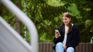 Nuori nainen katsoo hymyillen älypuhelinta. Taustalla on vihreitä puita.