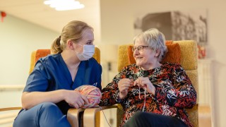 Hoitaja ja ikääntynyt nainen istuvat iloisina vierekkäin nojatuoleilla toisiaan katsoen.