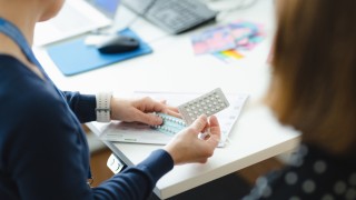 Kaksi henkilöä työpöydän ääressä, toisella kädessä lääkepakkaus.