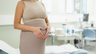 Raskaana oleva nainen muodostaa käsillään sydämen vatsansa päälle sairaalaympäristössä.