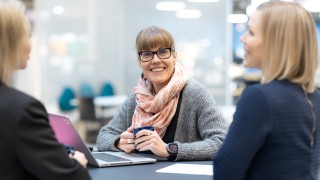 Kolme henkilöä istuu työpöydän ääerssä. Yksi heistä katsoo kameraan ja hymyilee.